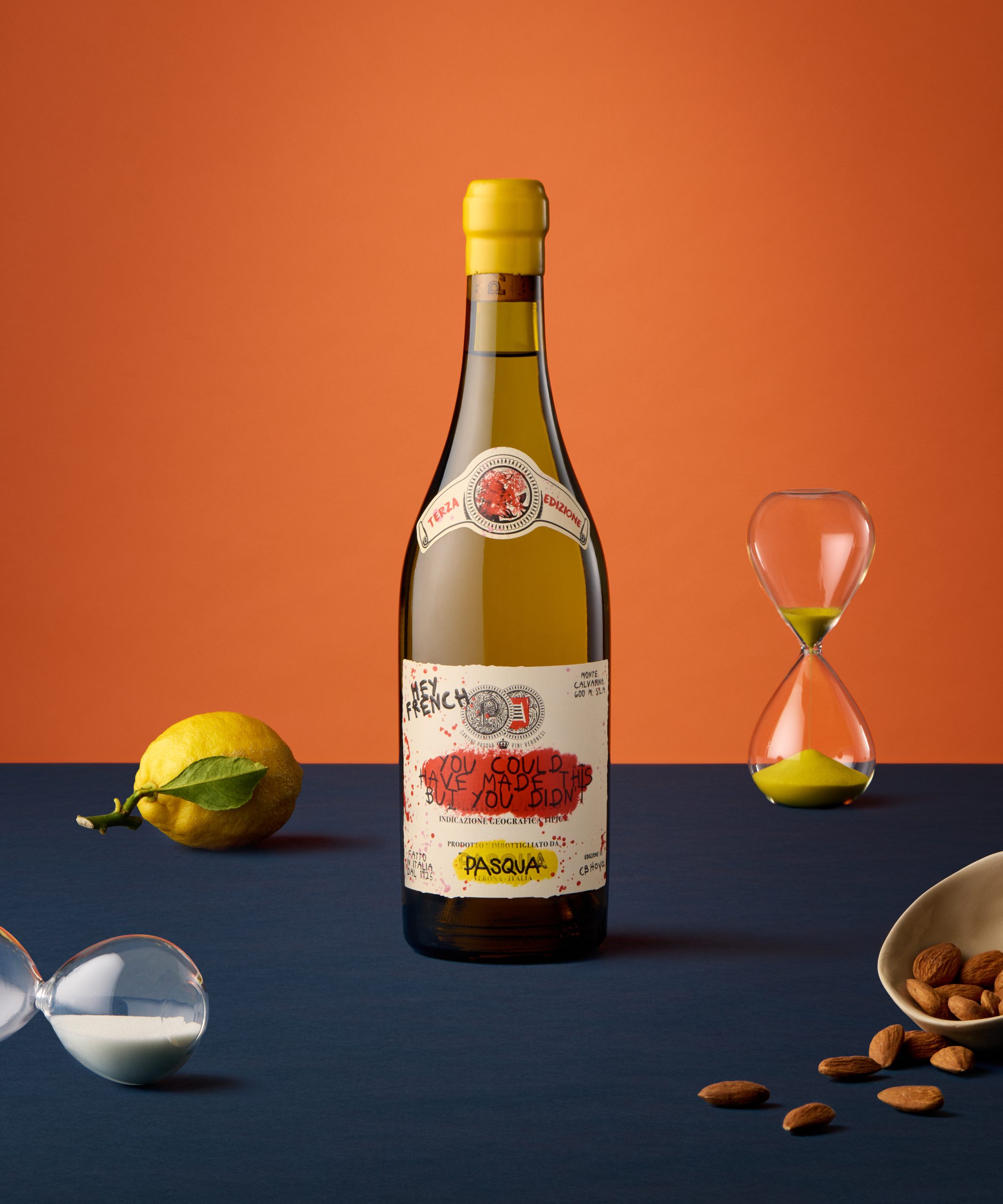 橙色背景的蓝色桌子上放着一瓶 "Hey French "葡萄酒。桌上还有沙漏、柠檬和杏仁。