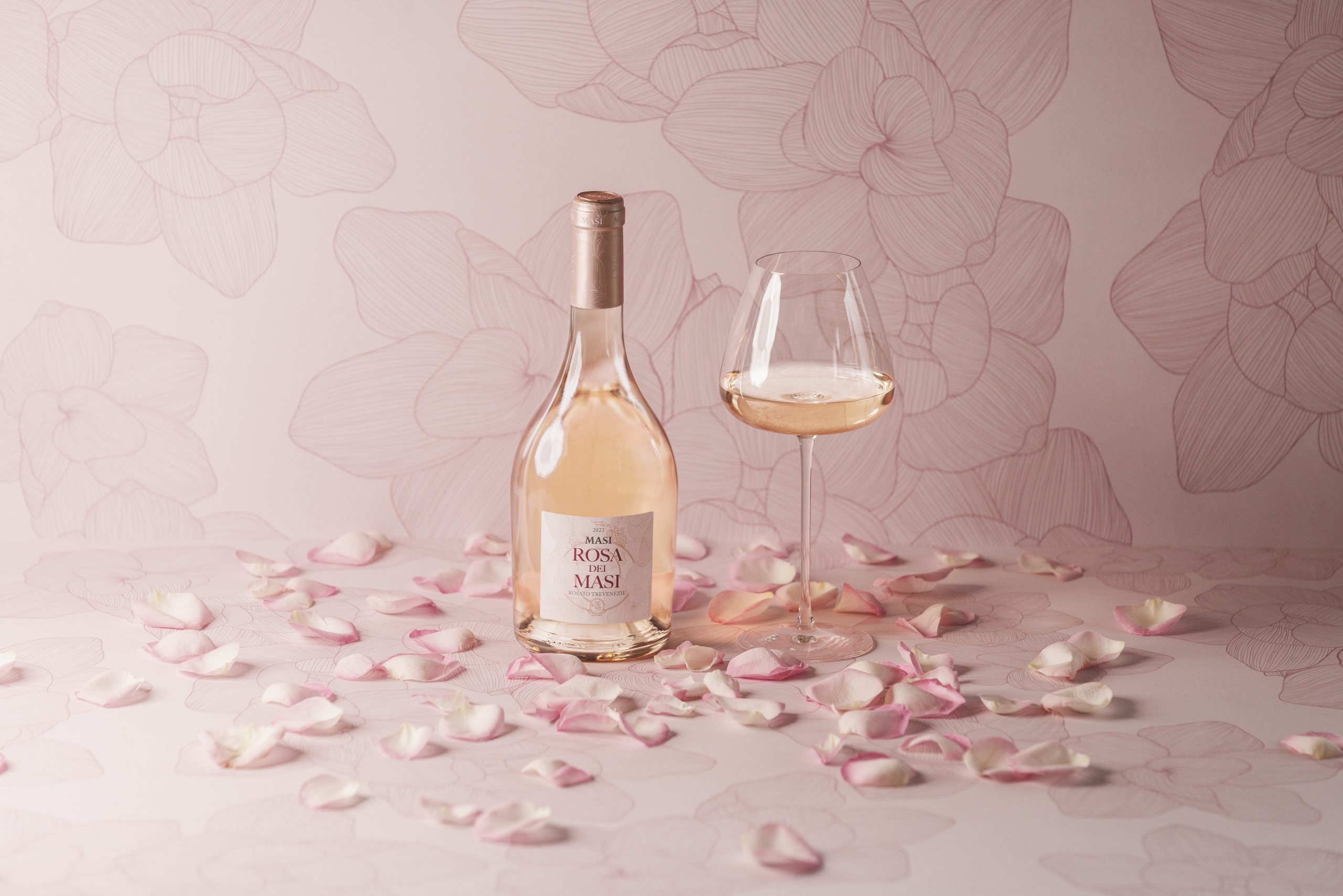 Una bottiglia del nuovo Rosa dei Masi si trova accanto a un bicchiere di vino. I petali di rosa sono sparsi in giro e il fondale rosa pallido presenta gli stessi motivi di rosa disegnati a inchiostro della bottiglia.