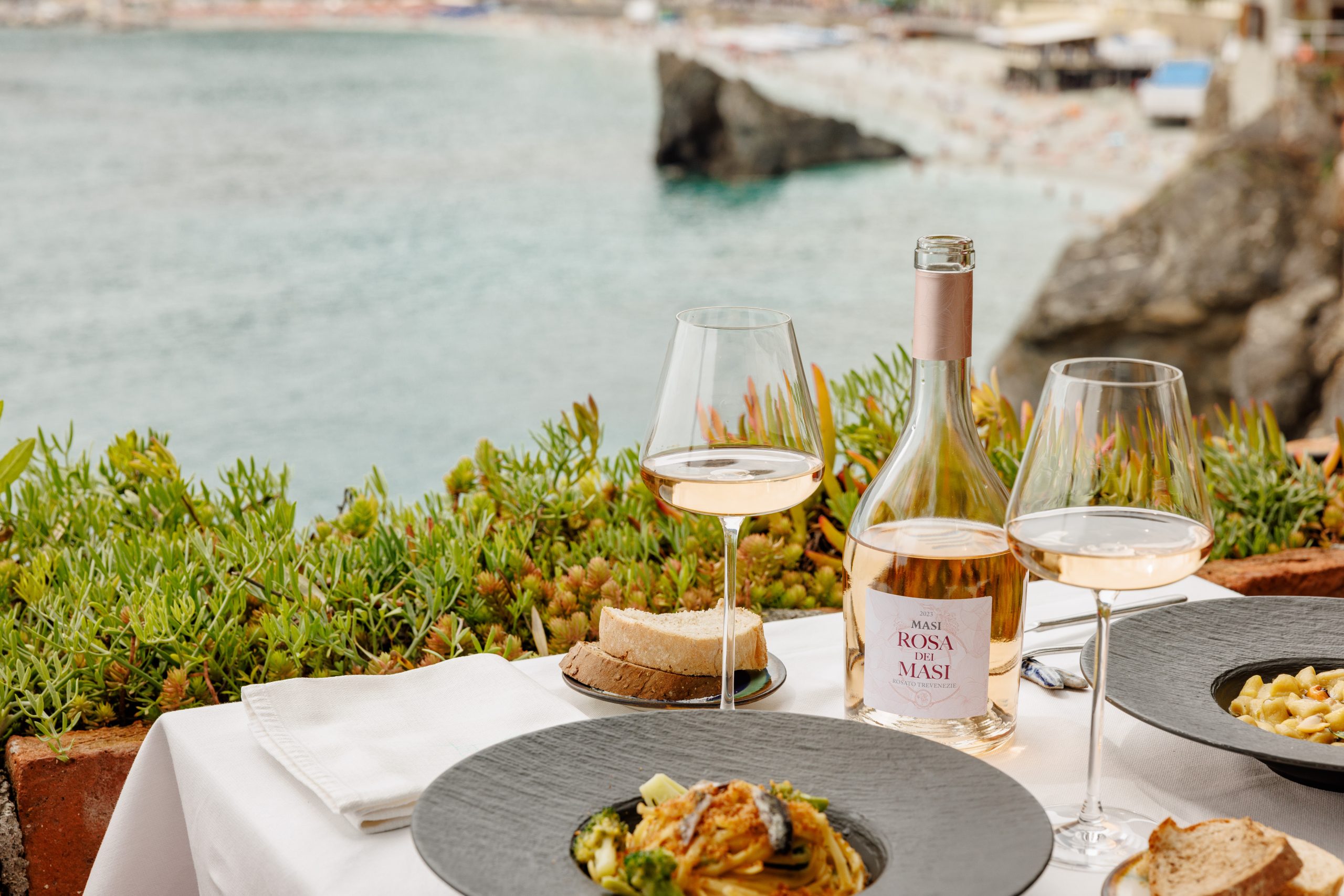 Un tavolo è apparecchiato per un pasto con vista su una baia. Su di essa si trovano una bottiglia di Rosa dei Masi, due bicchieri di vino, pane, un piatto di pasta e tovaglioli.