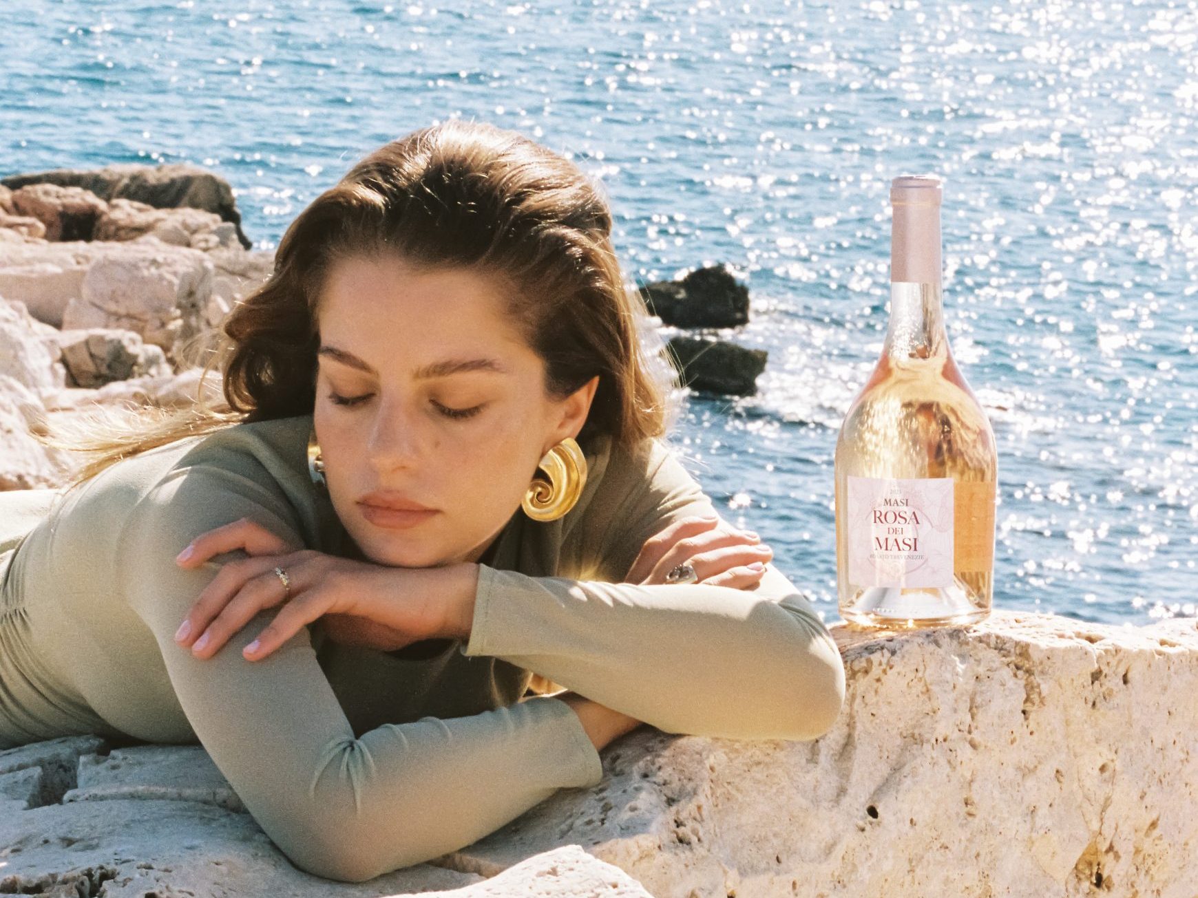 Une femme portant un haut vert se repose sur des rochers blancs au bord de la mer. Une bouteille de Rosa dei Masi est posée sur les rochers à côté d'elle.